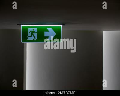 Green fire escape sign hang on the ceiling in the dark building. Emergency fire exit sign, warning plate with running man icon and arrow to the right Stock Photo