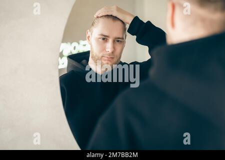 An attractive young man with a beard looks at himself in the mirror. Stock Photo