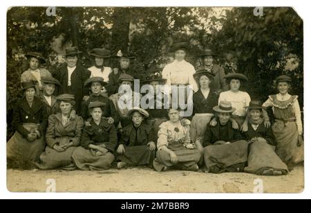 Original late Edwardian era postcard of group of working class women, lots of characters of different ages. Many are wearing the lady's version of the flat cap. Believed to be hop pickers as appear to be in a country setting, is a large group of different ages  and several are wearing posies of what look like a bunch of hops on their lapels and wearing  aprons tied at waist, over urban clothes. Many London workers went to work in the Kent hop fields on a working holiday.  Circa 1905. Stock Photo