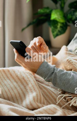 a caucasian man relaxing, using smart phone lying in bed under throw at home Stock Photo