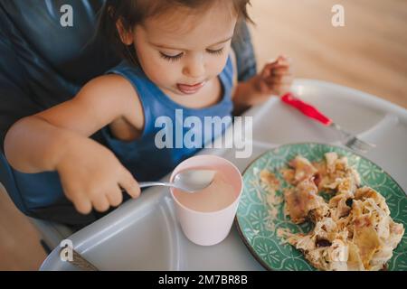 Cute baby girl eating food by herself, sitting on high chair. Caucasian infant self-feeding solid food fine motor development. Stock Photo