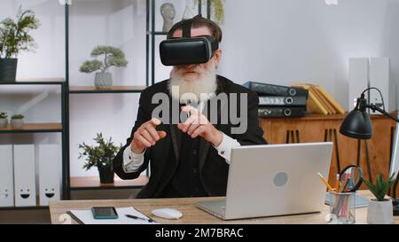 Senior businessman programmer software developer using headset helmet app to play simulation game. Watching virtual reality 3D 360 video. Senior old grandfather in VR goggles working at home office Stock Photo