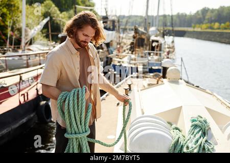 Portrait of handsome long haired man standing on boat in dock and holding line, lit by sunlight, copy space Stock Photo