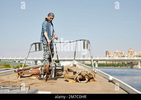 Full length portrait of tough young man standing on boat and tying rope while sailing in city waters, copy space Stock Photo