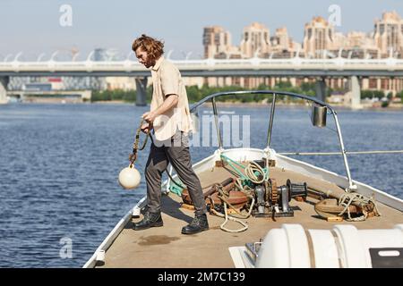 Full length side view of handsome young man standing on boat and throwing line in water, copy space Stock Photo