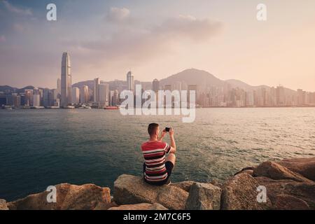 Rear view of man photographing Hong Kong urban skyline at sunset. Tourist using smart phone in modern city. Stock Photo