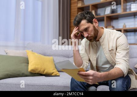 Upset man at home sitting on sofa reading letter with bad news notification in envelope opening, unhappy in living room and sad. Stock Photo