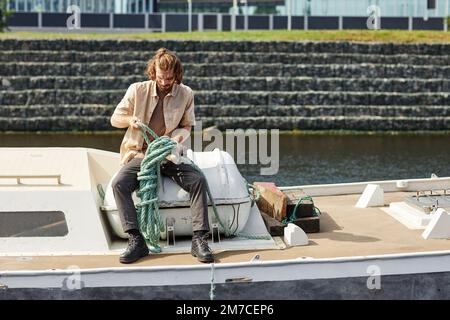 Full length portrait of long haired man on boat tying rope in sunlight, copy space Stock Photo