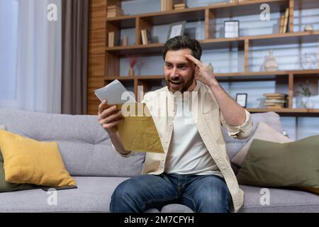 Joyful man at home holding envelope with notification message reading and smiling happy with news sitting on sofa inside living room. Stock Photo
