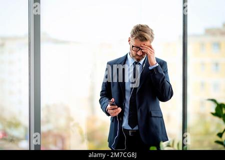 Thoughtful male professional using smartphone and earphones while standing at the office. Professional man wearing glasses and suit. Stock Photo