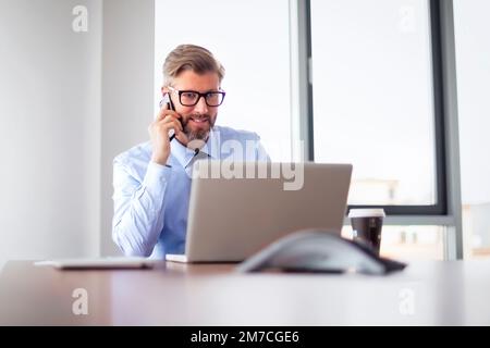 Mid adult business professional sitting at table with laptop and using mobile phone. Thinking businessman working in office. Stock Photo