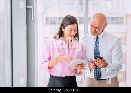 Business people standing together at the office and using digital tablet and smartphone.. Attractive businesswoman and middle aged businessman discuss Stock Photo
