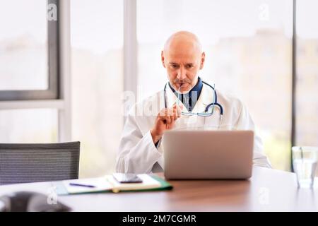Shot of a mature doctor sitting alone in his clinic and looking contemplative while using his laptop. Stock Photo