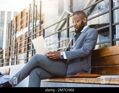 A dark-skinned man in a business suit is using a laptop outdoors. Digital education and remote work Stock Photo