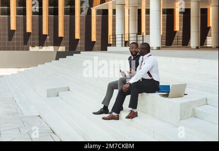 Two African American businessmen in suits communicate. Friends and business partners sit on the concrete steps of the city stairs against the backdrop Stock Photo