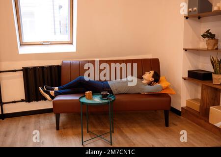 Serene housewife lady, living room, sitting on sofa, feeling tired, holding hands behind head, calm young woman resting on comfortable sofa, breathing Stock Photo