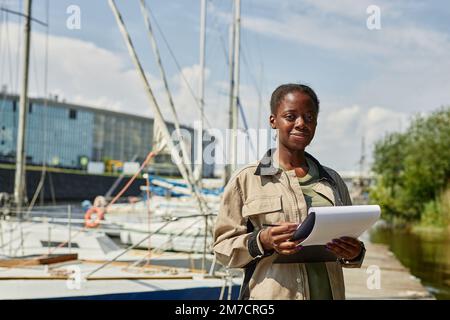 Waist up portrait of black young woman holding clipboard and smiling at camera while working in yacht docks, copy space Stock Photo