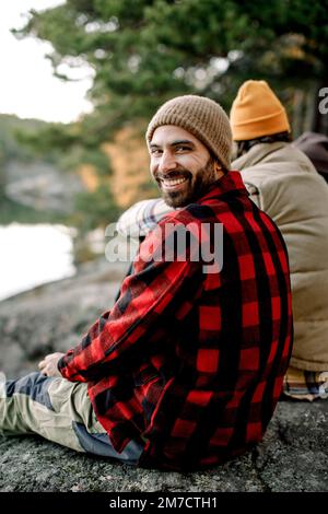 Happy man wearing plaid shirt looking over shoulder while sitting on rock Stock Photo