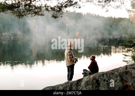 Male friends talking to each other on rock near lake during staycation Stock Photo