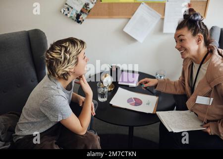 Smiling non-binary psychologist looking at teenage student discussing while sitting in school office Stock Photo