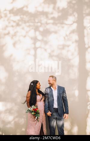 Happy multiracial bride and groom holding hands against wall Stock Photo
