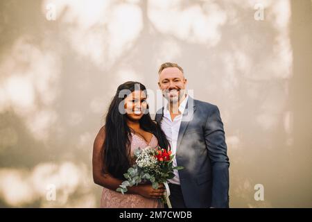 Portrait of smiling bride holding bouquet standing by groom against wall Stock Photo