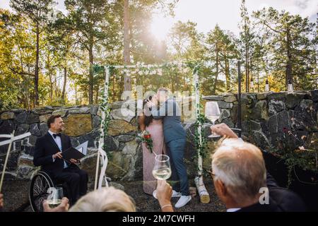 Newlywed couple kissing in front of guests raising toasts and minister with disability at wedding ceremony Stock Photo