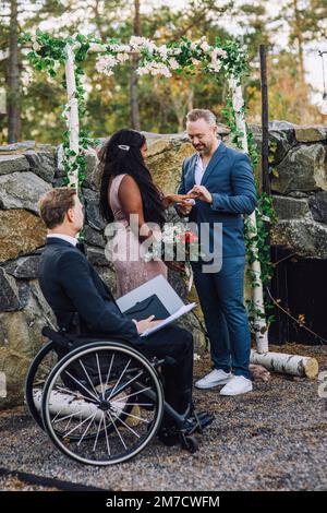 Smiling groom and bride exchanging ring by minister in wheelchair with disability at wedding ceremony Stock Photo