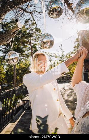 Happy groom holding bride's hand dancing under disco balls on sunny day Stock Photo