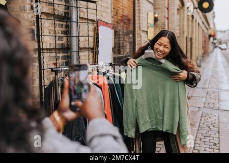 Mature woman photographing cheerful female friend trying on clothes at street Stock Photo