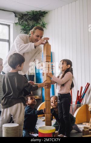 Male teacher guiding children stacking wooden toy blocks at kindergarten Stock Photo
