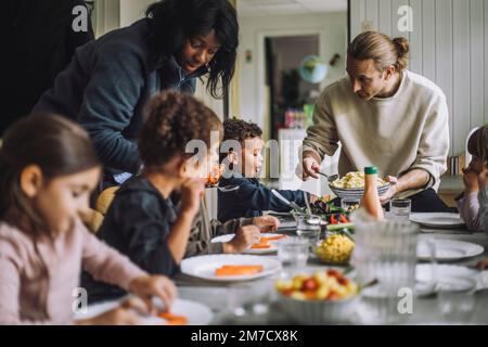 Male and female teacher serving food to children while having meal at table in kindergarten Stock Photo