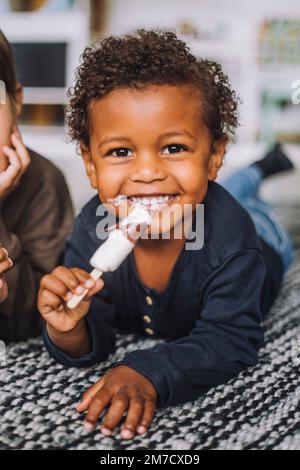 Portrait of happy boy having ice cream by male classmate lying on carpet in kindergarten Stock Photo