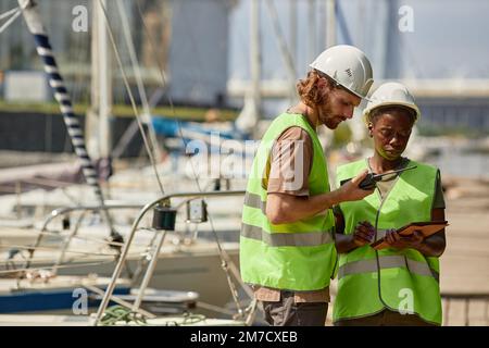 Waist up portrait of two young workers wearing hardhats while standing in yacht docks lit by sunlight, copy space Stock Photo