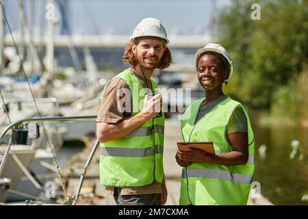 Waist up portrait of two young workers wearing hardhats and smiling at camera while standing in yacht docks Stock Photo