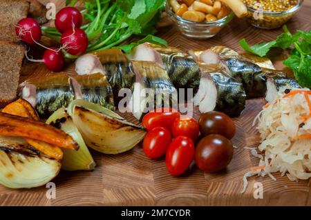 Smoked mackerel fish with vegetables on wooden board with appetizers: cherry tomato, baked onion and carrot, sauerkraut, mushrooms, radish. Stock Photo