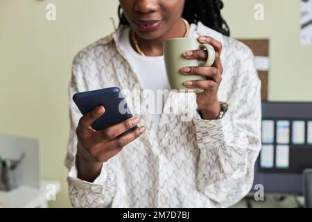 Close up of young black woman using smartphone and enjoying coffee while taking break from work, copy space Stock Photo