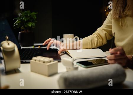 Woman sitting at table in dark room and using laptop. Working late from home office. Female freelancer works in home office at night Stock Photo