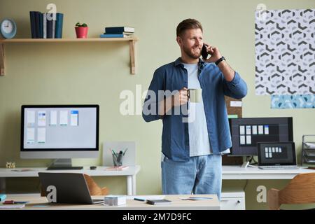 Portrait of smiling young man calling by phone and enjoying coffee while taking break from work in office, copy space Stock Photo