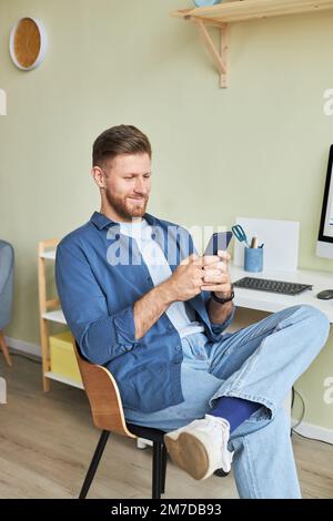 Vertical portrait of smiling young man relaxing at workplace and using smartphone Stock Photo