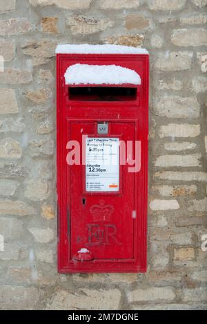 A deep red post box set in a wall in teh Cotswolds, covered in winter ...