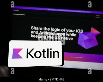 Person holding mobile phone with logo of programming language Kotlin on screen in front of web page. Focus on phone display. Stock Photo