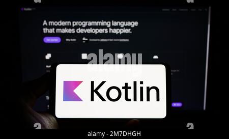 Person holding cellphone with logo of programming language Kotlin on screen in front of webpage. Focus on phone display. Stock Photo