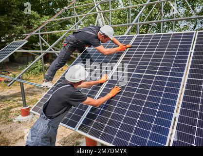 Workers installing photovoltaic solar panel system outdoors. Men engineers placing solar module on metal rails, wearing construction helmets and work gloves. Renewable and ecological energy. Stock Photo
