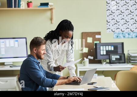 Side view portrait of two smiling young colleagues working together and using laptop in development agency office, copy space Stock Photo