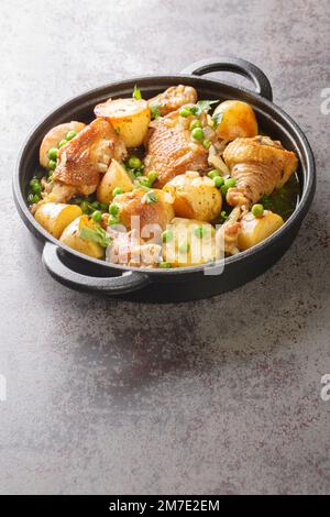 Delicious pieces of chicken cooked with potatoes and green peas close-up in a frying pan on the table. Vertical Stock Photo