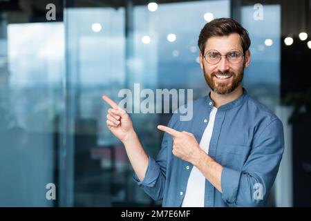 Smiling businessman inside office pointing fingers to direction, happy boss in shirt and glasses. Stock Photo
