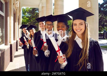 Multinational group of attractive students-graduate in mortar boards cap and ceremonial robes holding diploma in their hands. Stock Photo