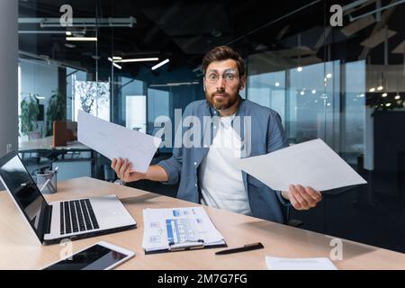 Dissatisfied and angry businessman inside office showing reports and bills documents to camera, boss in shirt looking at camera working with laptop paper work. Stock Photo