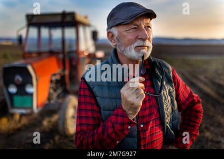 Pensive mature farmer standing in front of tractor in field in autumn Stock Photo